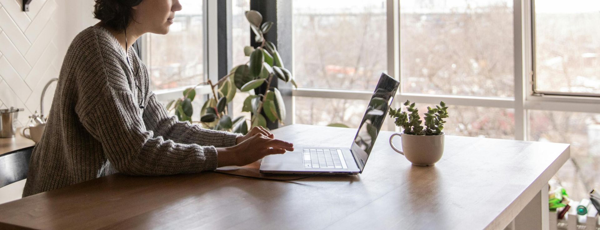 Side view of young female in casual clothes typing message on laptop while sitting at wooden table in kitchen