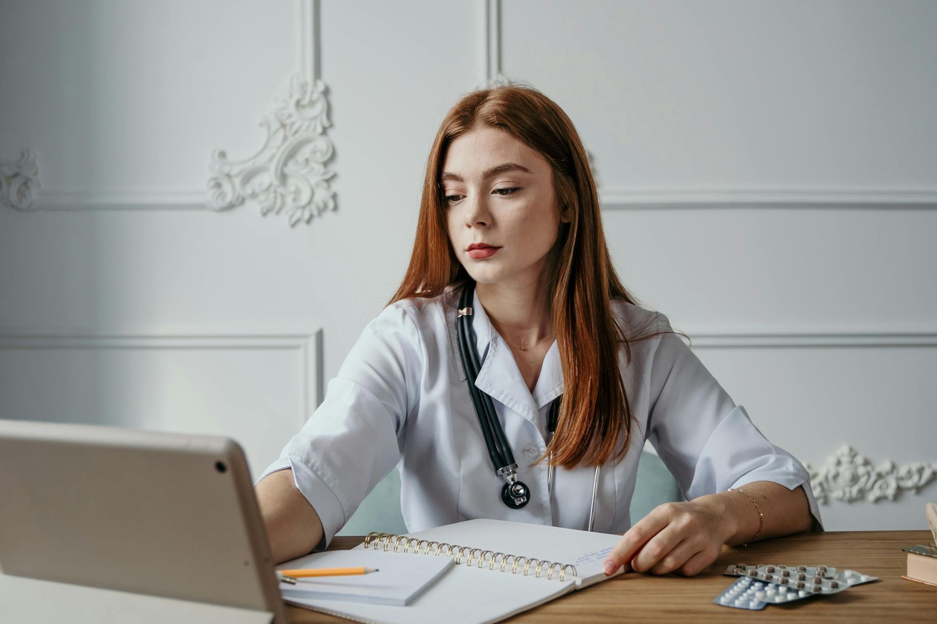 A professional female doctor wearing a stethoscope, working on a laptop at a desk with notebooks and medicines.